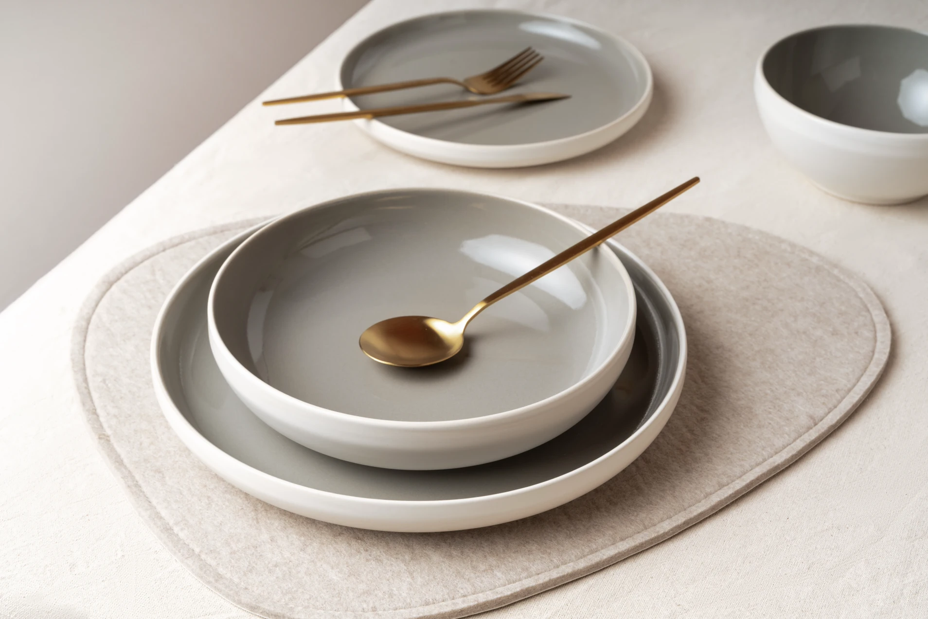Gedeckter Tisch mit CAVO Teller und Schale in Stone auf einem Platzset aus Filz, dazu goldenes Besteck